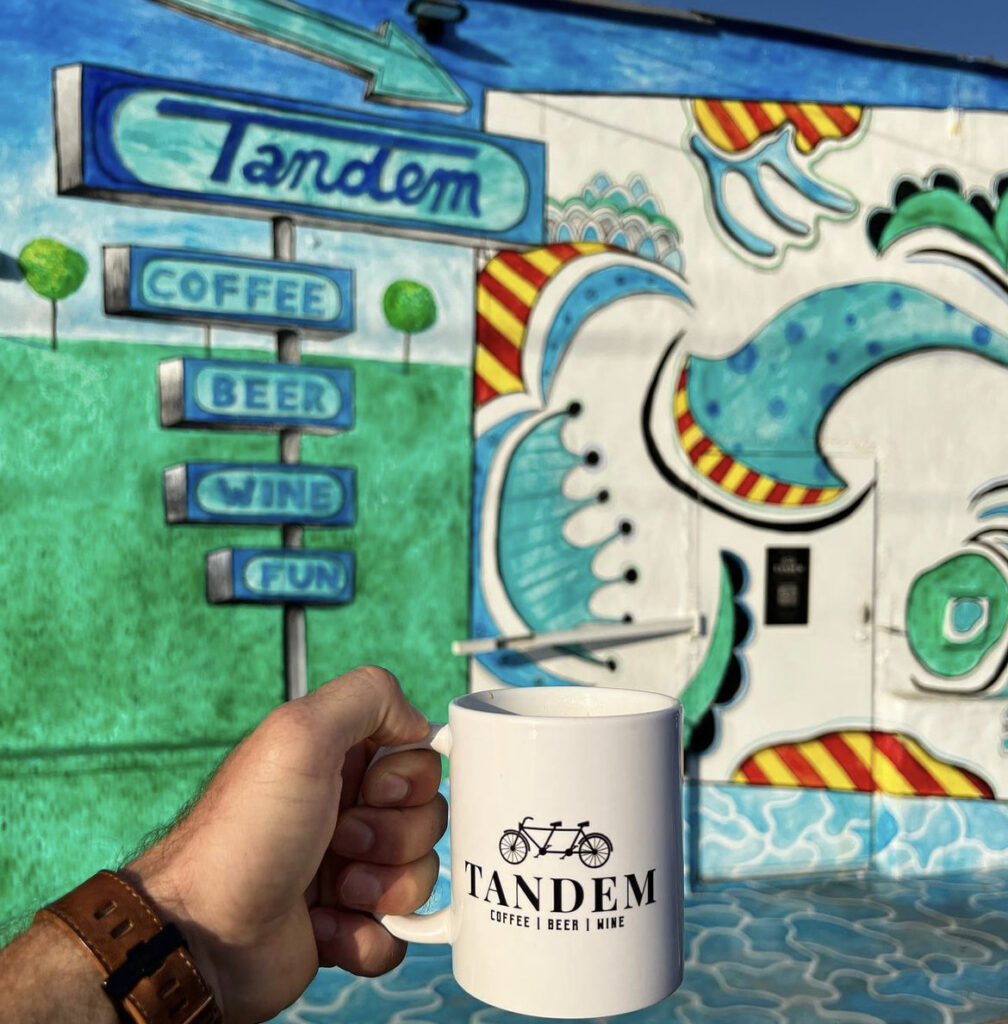 A coffee mug with the tandem logo appears in front of colorful coffee shop in San Antonio.