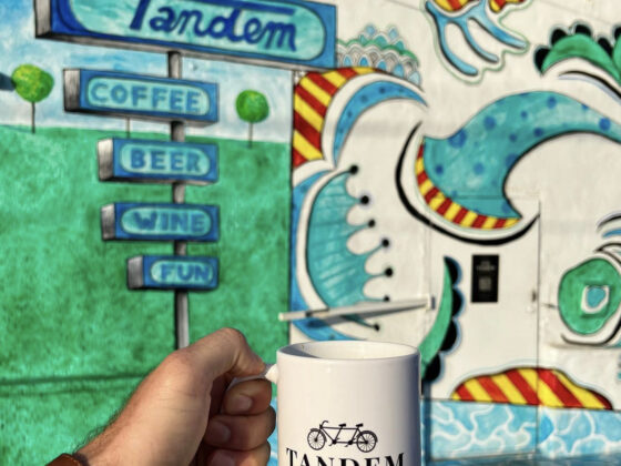 A coffee mug with the tandem logo appears in front of colorful coffee shop in San Antonio.