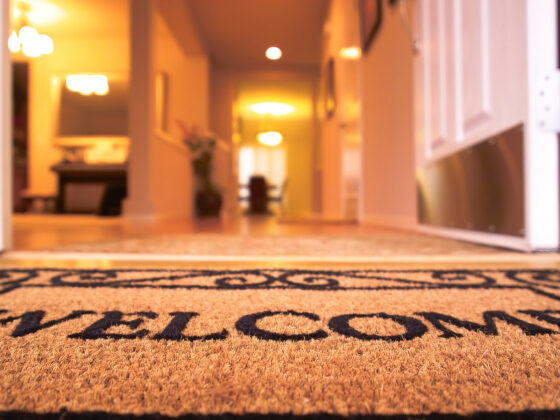 From a low angle, a burlap welcome mat lies at the door of a home with warm blurry light in the background