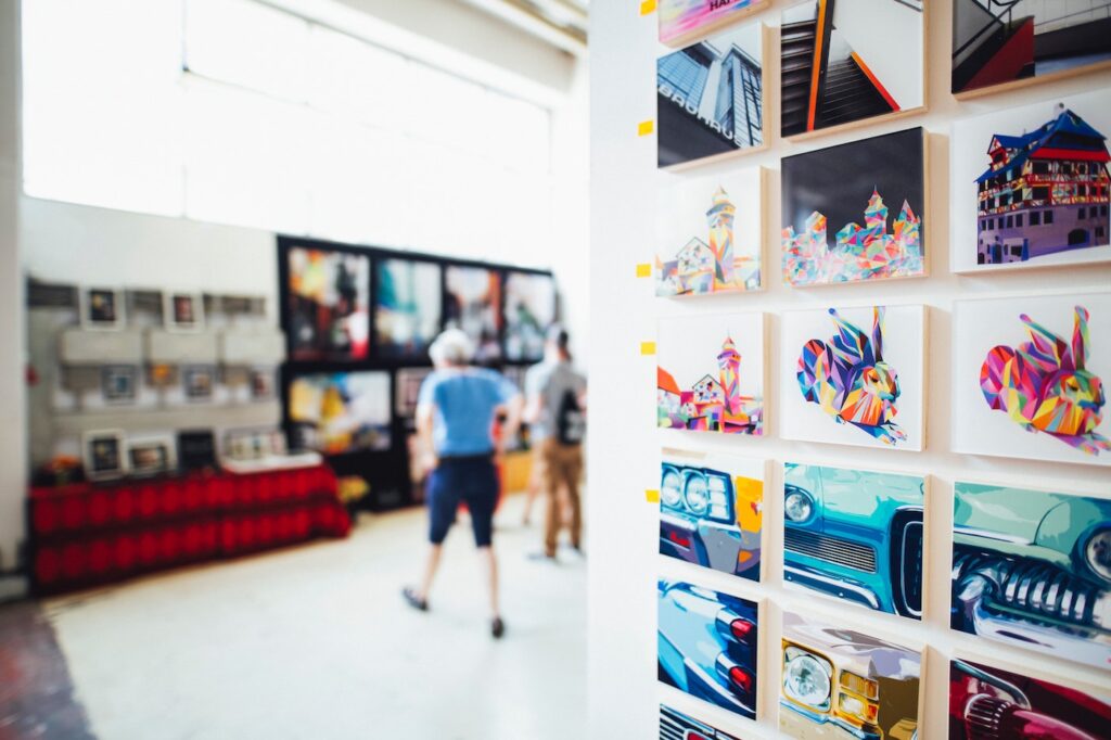 the interior of an an art gallery with multiple pieces of art on the wall, and two people in the background looking at art.