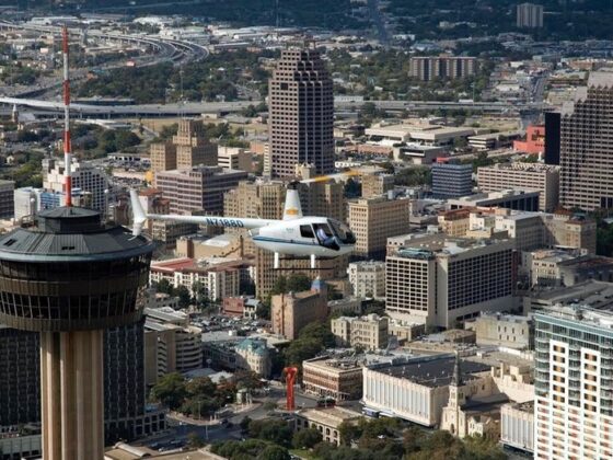 A helicopter flies high over the San Antonio skyline with the Tower of the Americas nearby.