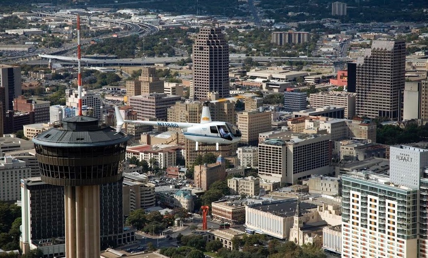 A helicopter flies high over the San Antonio skyline with the Tower of the Americas nearby.