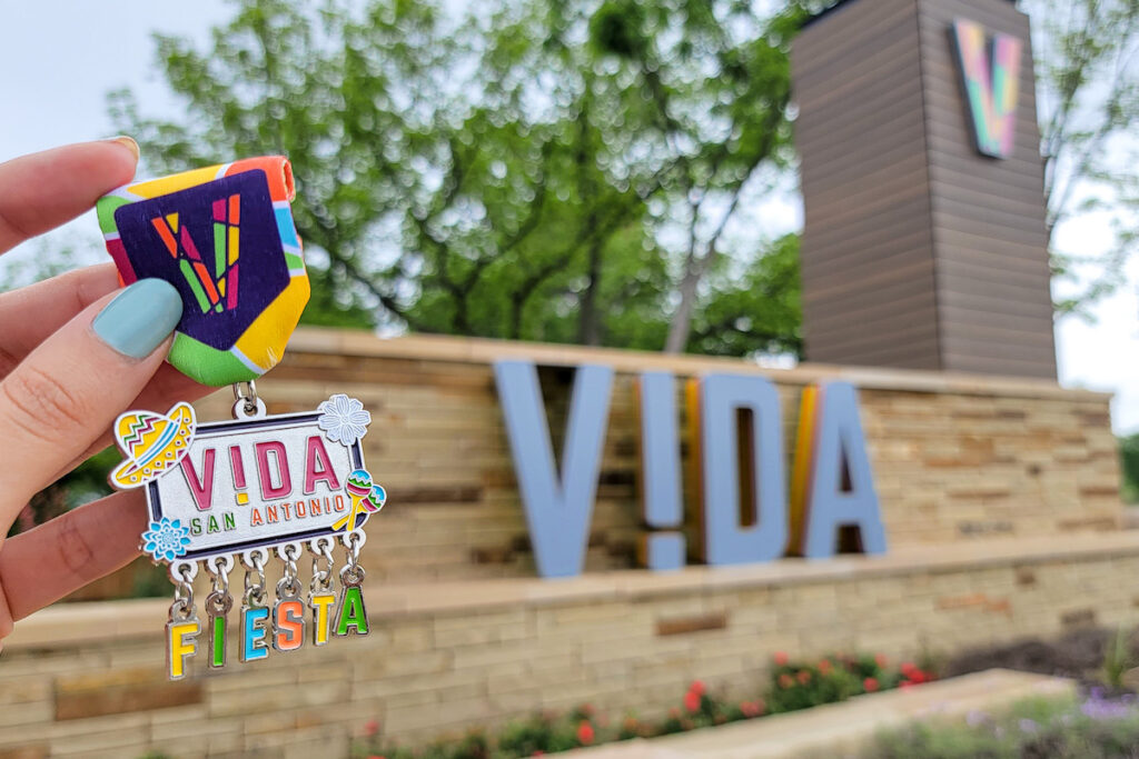 A VIDA San Antonio 2023 Medal is held up against the VIDA entrance side on the Southside.