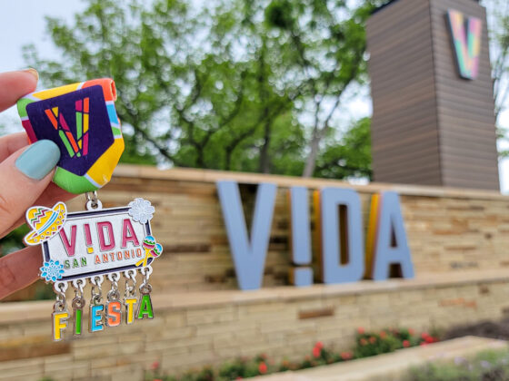 A VIDA San Antonio 2023 Medal is held up against the VIDA entrance side on the Southside.