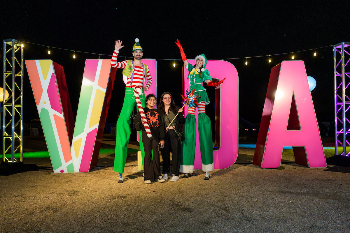 A family poses with festive stilt walkers in front of the VIDA sign