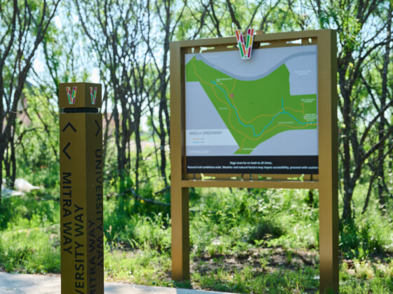 A signpost marked "Mitra Way" stands beside a map board under trees. The map depicts a section of the Madla Greenway with pathways and a "You Are Here" indicator amidst green foliage on this bright, sunny day.