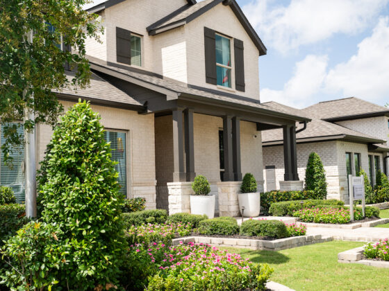 A suburban house with beige brick, dark trim, and a covered porch in the VIDA San Antonio community. The front yard boasts manicured bushes, flower beds, and a small tree. A real estate sign is visible near the walkway under the partly cloudy sky.