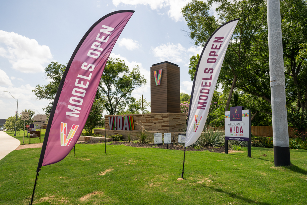 "Model Homes Now Open" flags wave in front of colorful VIDA San Antonio signage at the community entrance.