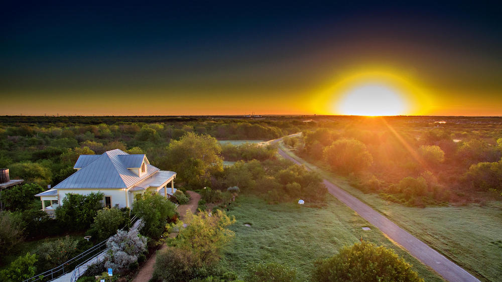 A sunrise against the horizon above the Mitchell Lake Audubon Entry building, sprawling vegetation around