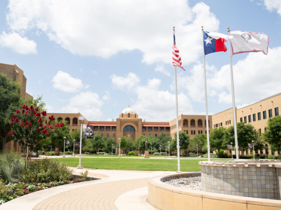 The American flag, the Texas Flag, and the Texas A&M flag stand waving in front of the Texas A&M San Antonio campus