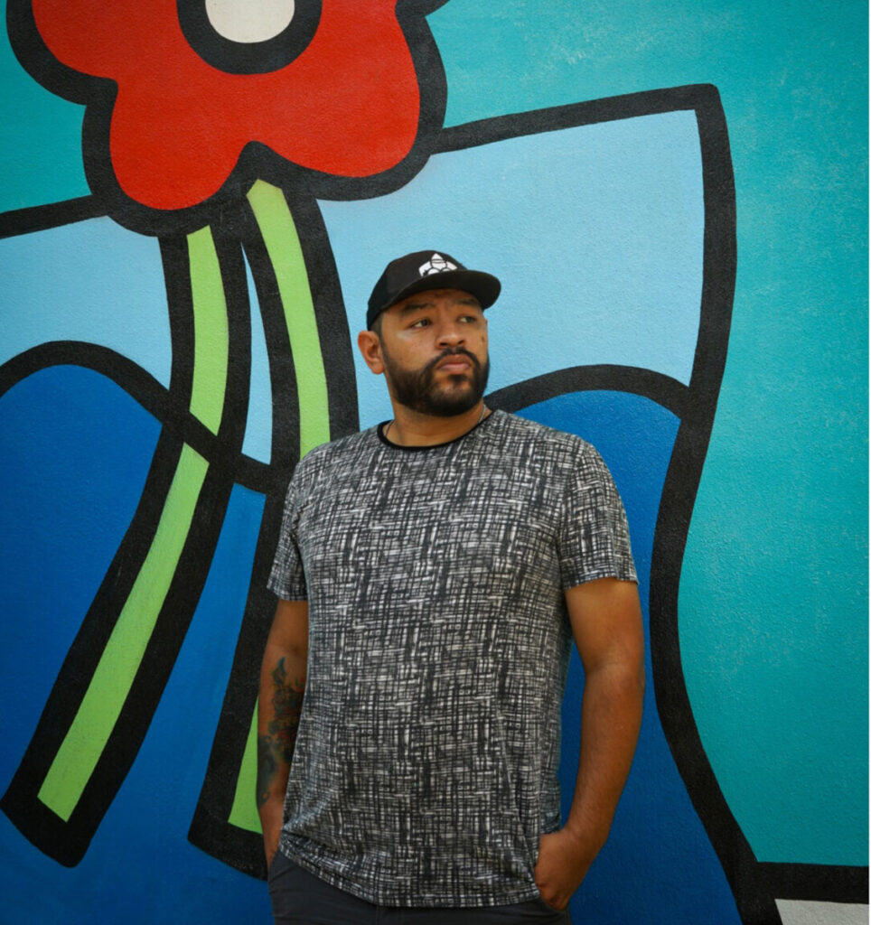 Albert Gonzalez, Trebla Art, stands against a larger than life painting of his that features a wabi-sabi flower, in primary colors with thick black lines.