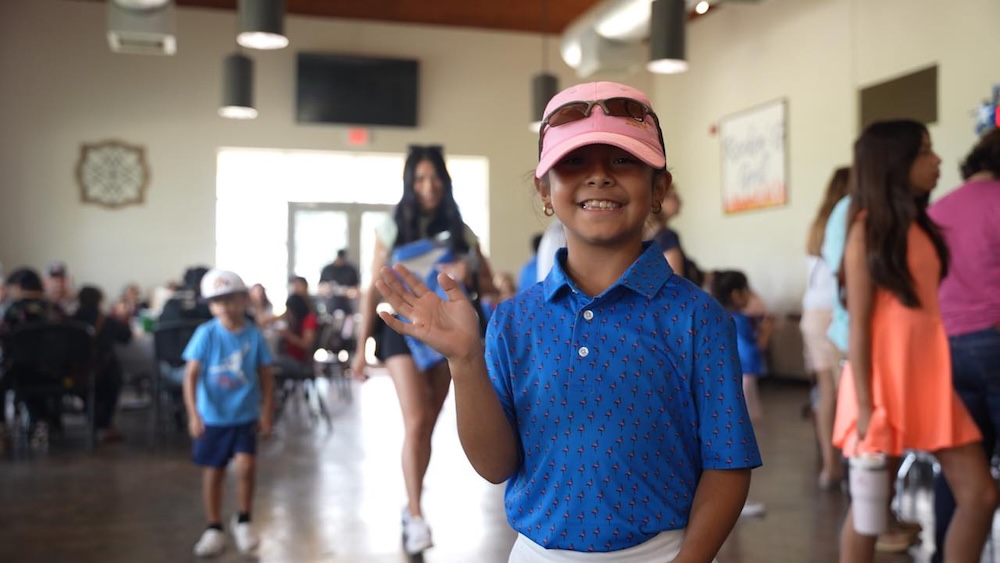 A young golfer in a pink baseball cap waves