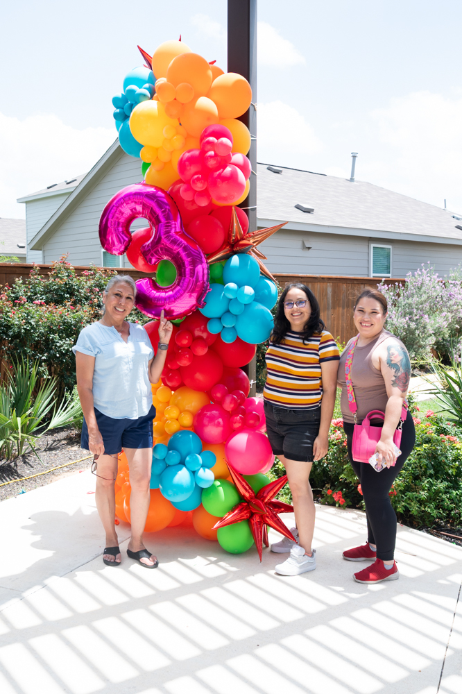 Three women stand around a balloon centerpiece with colorful balloons, on in the shape of a large number "3"