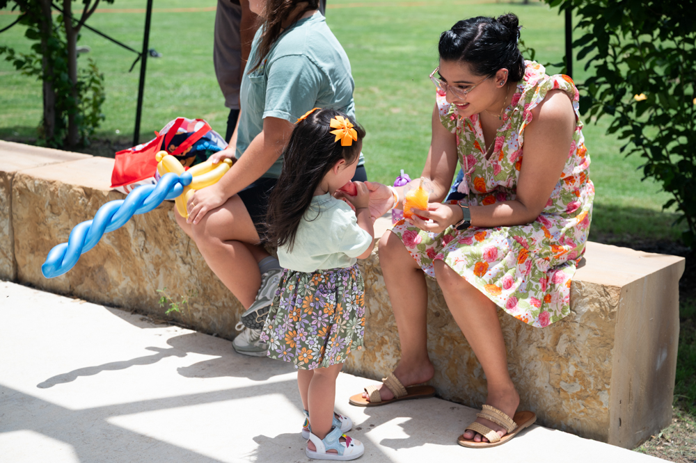 A mom on a stone bench leans over to talk to her toddler on a sunny day
