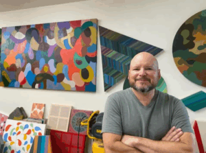 Artist Louis Vega Trevino poses for a photo in his studio, art on the walls behind him