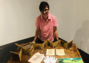 Artist Sarah Booth stands against a table with art laying out on it