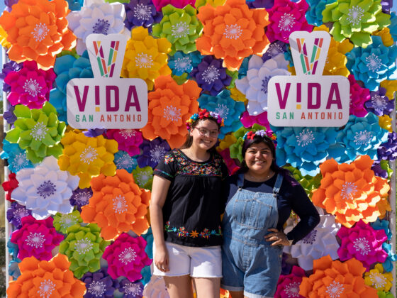 Two women in flower crowns celebrate San Antonio Fiesta in front of a brightly colored paper flower wall with VIDA San Antonio community logos