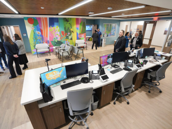 The inside of the Behavioral Health section of the University Health Vida clinic. The room is large and bright, with acoustic tiles on the ceilings, a colorful flower mural on the wall, and a long reception desk with three computers and chairs. Several visitors can be seen conversing with each other in the background