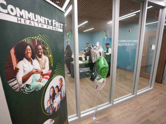A large green banner reading "Community First Health Plans" can be seen outside of a room labeled "Community First Health Plans." The walls are made of large windows so you can inside to the room, which has blue walls and a table with a greeter