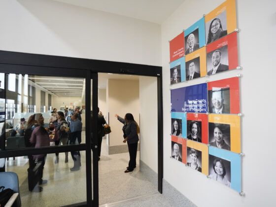 A wall with orange, blue, and yellow color blocks and several photos of the Bexar County Commissioners Court. Sliding doors can be seen next to the wall with several people touring the inside of the University Health Vida clinic