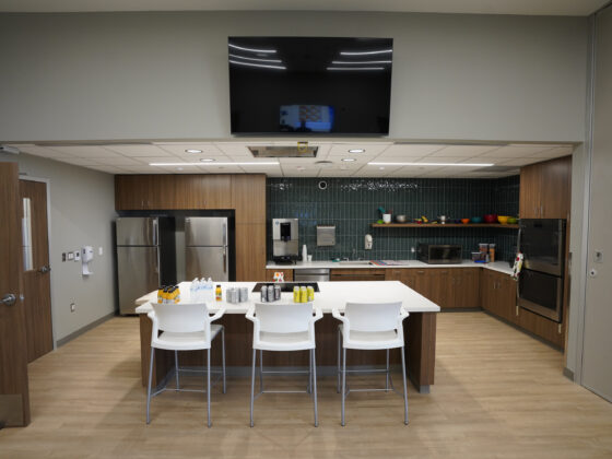 Kitchen inside the University Health Vida clinic. A large kitchen island with three chairs are seen in the center, with two refrigerators in the background, a sink, colorful blue backsplash subway tiles, and cabinets. Bowls and pots can be seen on a floating shelf above the sink.