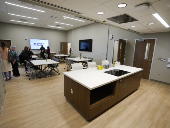 Large kitchen room inside the University Health Vida clinic. A kitchen island with a burner and several cabinets can be seen in the foreground with tables in the background and a group of people