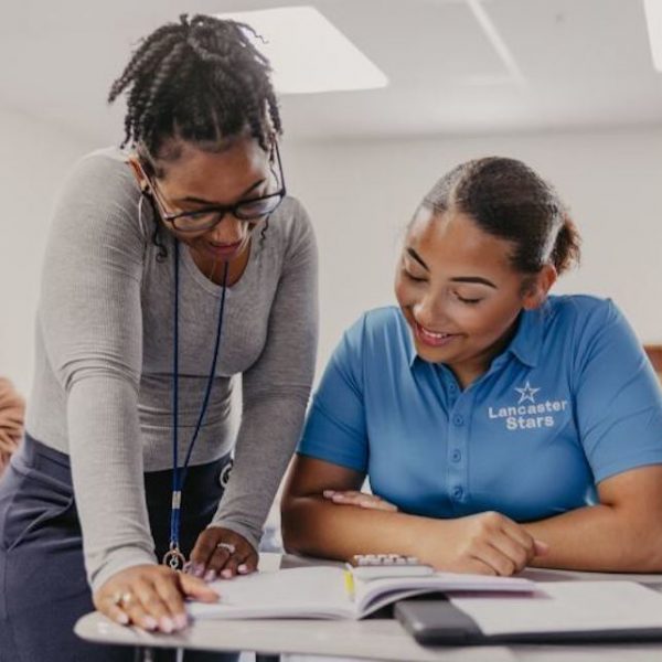 In a classroom near VIDA San Antonio, a teacher leans over a desk, smiling and pointing at a book. A student in a blue "Lancaster Stars" polo shirt looks on, grinning. Background chatter from other eager students fills the lively scene.