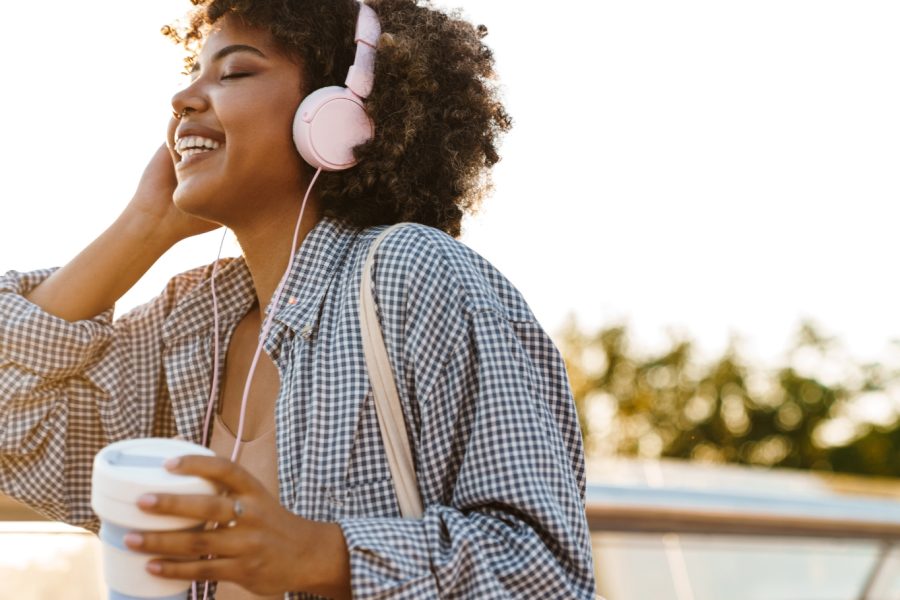 Curly haired young woman listens to music on pink headphones while walking through the town center and drinking a to-go coffee.